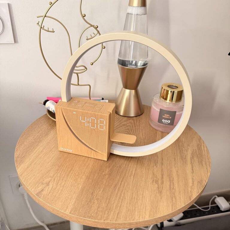 Le réveil-lampe est posé sur une table de chevet en bois clair, allumé, dans une chambre. On y voit l'heure 04:08.