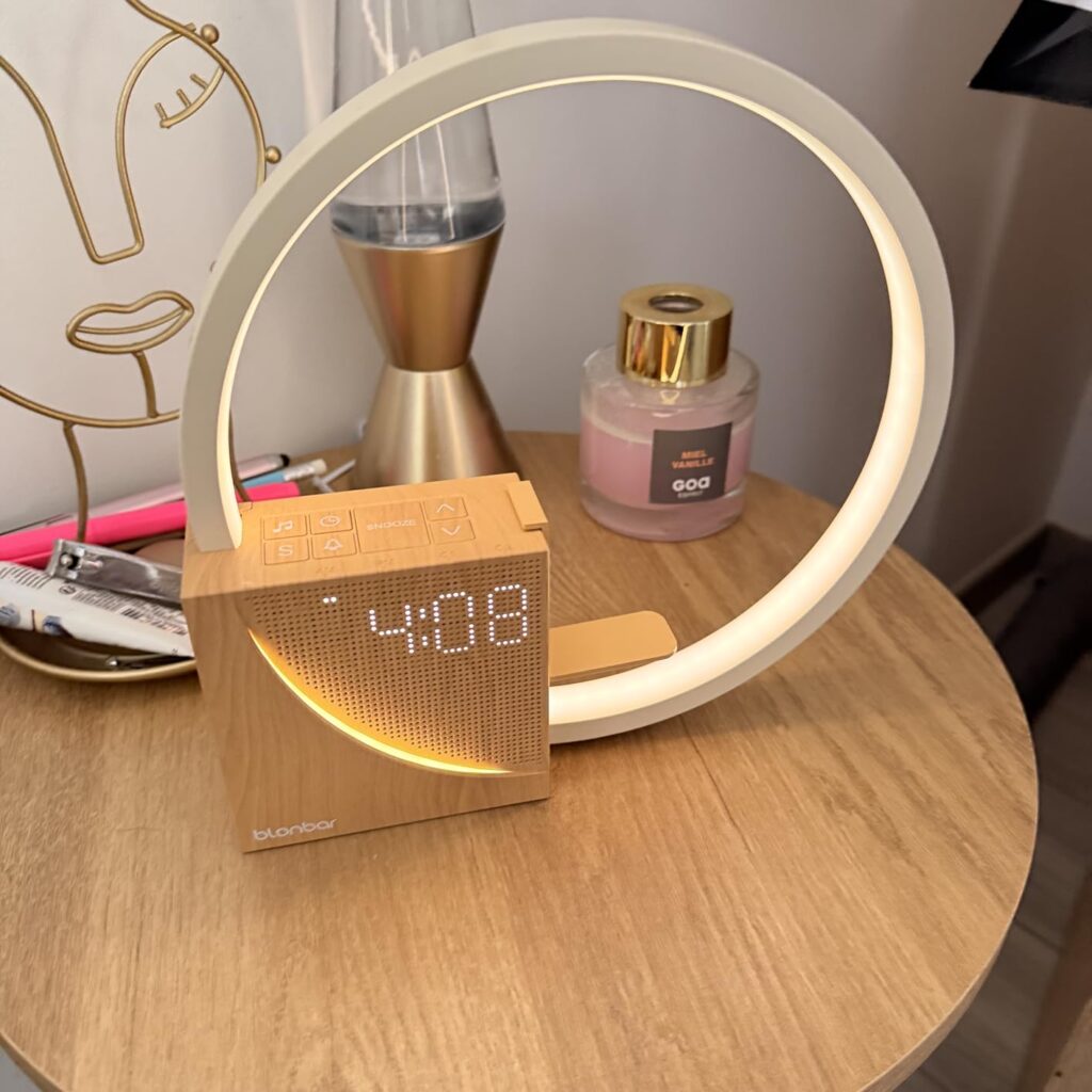 Le réveil-lampe est posé sur une table de chevet en bois clair, allumé, dans une chambre. On y voit l'heure 04:08. L'éclairag...