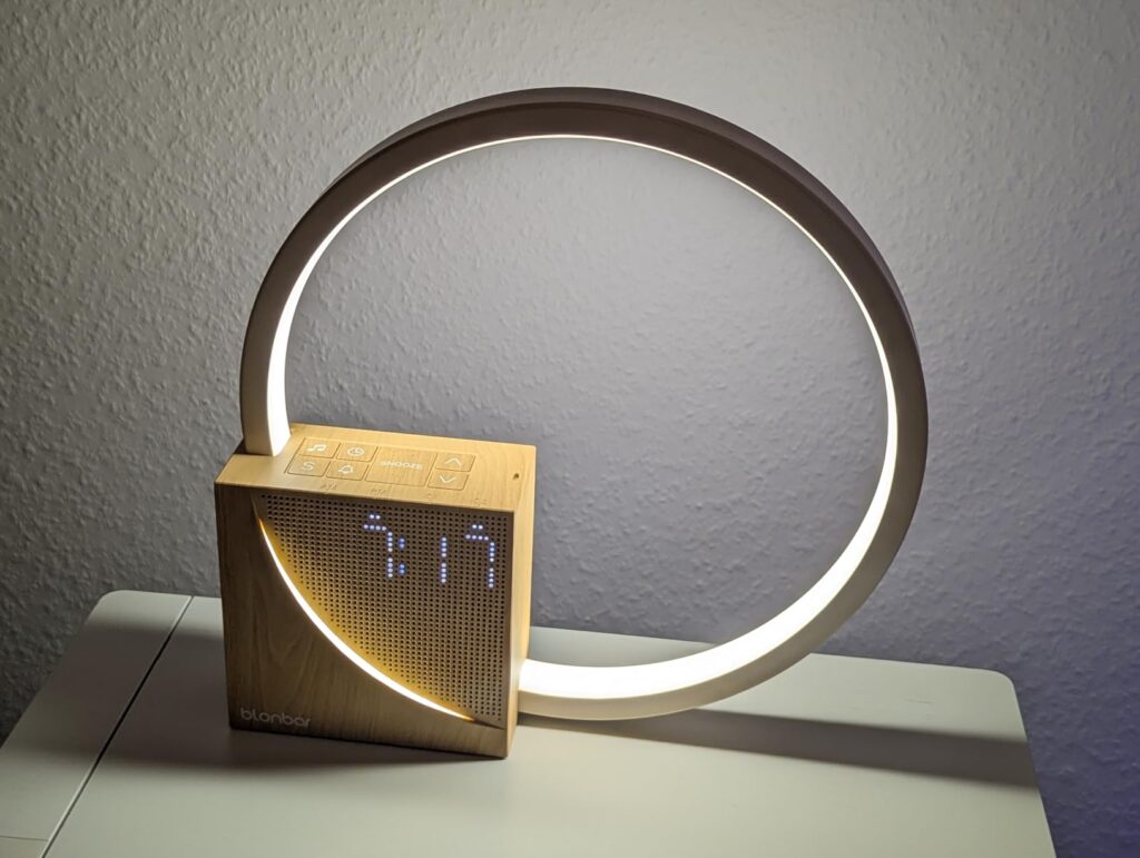 Le réveil-lampe est posé sur un meuble blanc, allumé, dans une pièce sombre. On y voit l'heure 7:17.