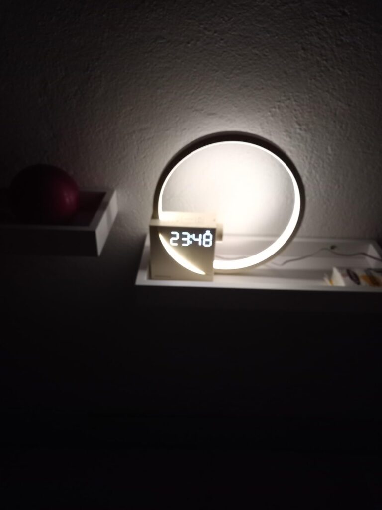 Le réveil-lampe est posé sur une étagère dans une pièce sombre. La lumière de l'appareil est allumée et l'heure est affichée.