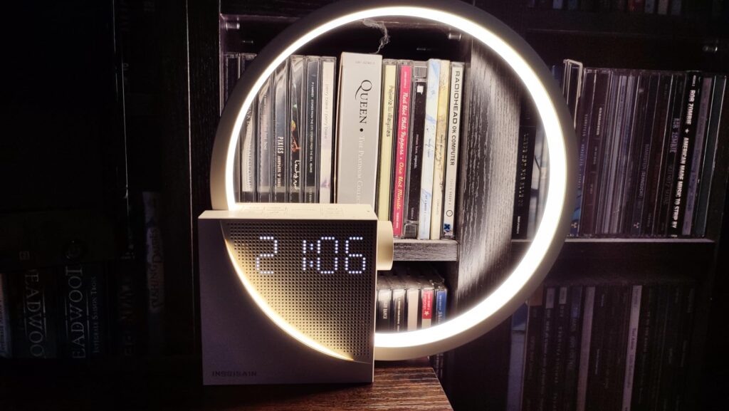 Le réveil-lampe est posé devant une étagère de livres, dans une pièce sombre. La lumière est allumée et l'heure est affichée.