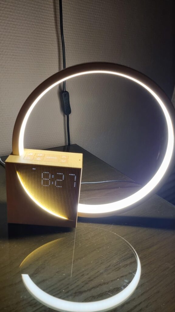 Le réveil-lampe est posé sur une table en bois, la lumière est allumée et l'heure est affichée. On aperçoit un câble à l'arri...