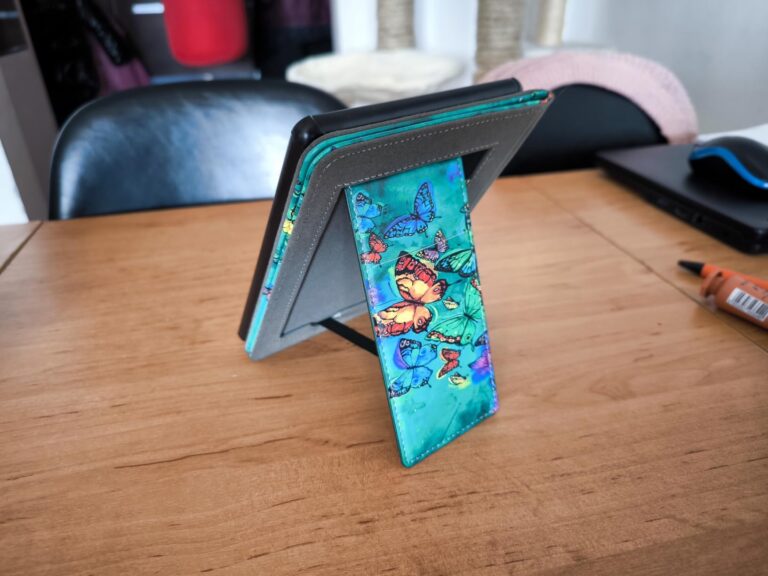 L'étui de liseuse est posé sur une table en bois, ouvert et servant de support pour la liseuse. Le motif de papillons est vis...
