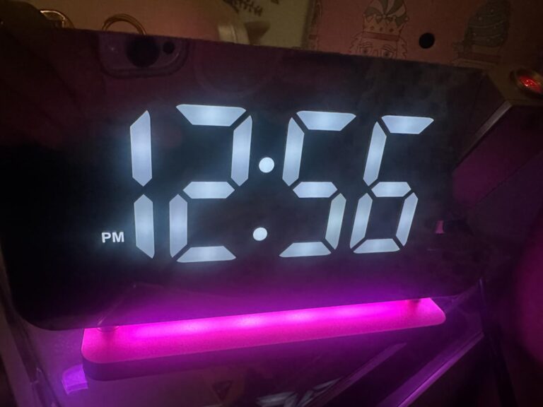Réveil en gros plan, affichant l'heure 12:56 et la mention 'PM', avec un éclairage d'ambiance rose.