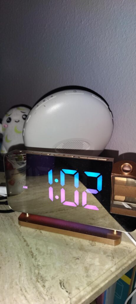 Réveil posé sur une table de nuit, affichant l'heure 1:02 AM avec des chiffres bicolores. L'éclairage d'ambiance est multicol...