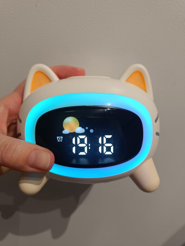 Vue frontale du réveil en forme de chat tenu dans une main. Les oreilles s'éclairent en orange et l'écran est entouré de bleu...