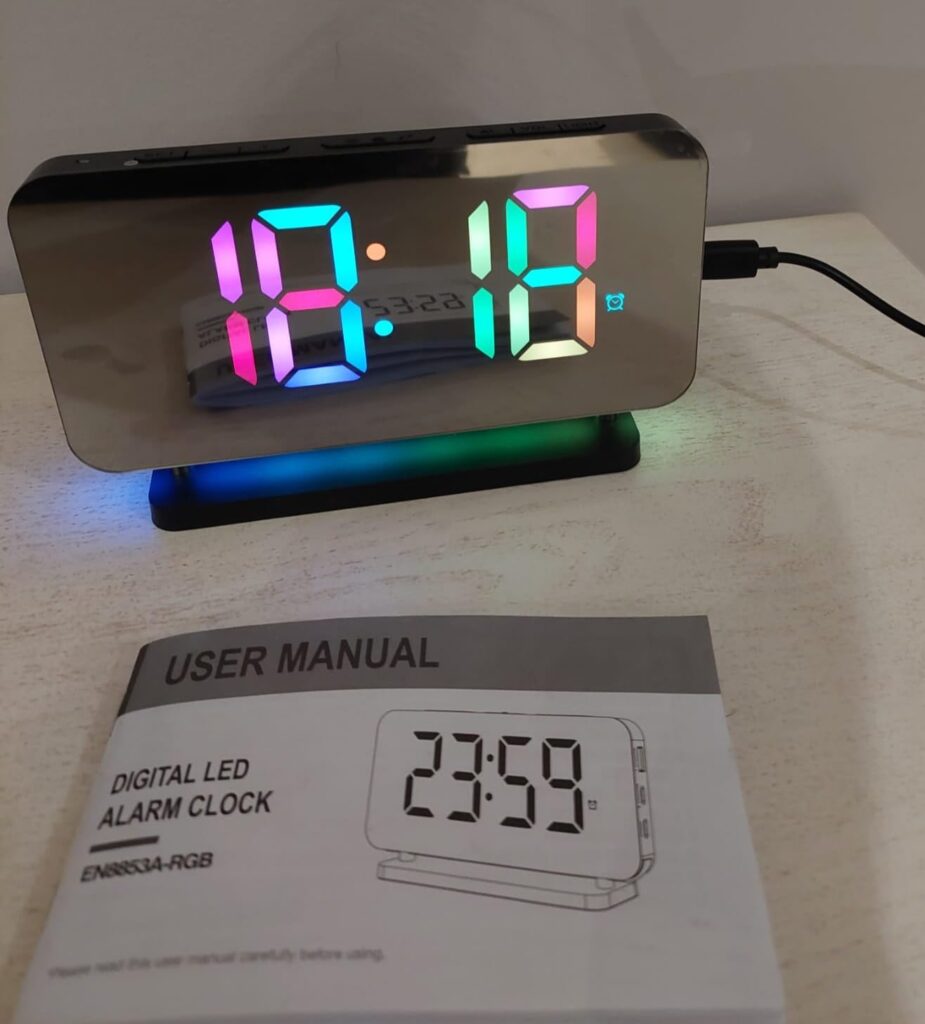 Le radio-réveil sur une table avec le manuel d'utilisation à côté. L'affichage est multicolore. Le manuel est ouvert à la pag...