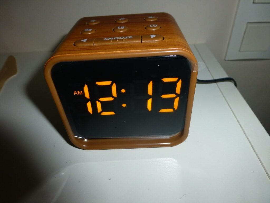 Le réveil est posé sur une surface blanche, le jour. L'heure est 12h13 AM, l'affichage est orange. Un câble d'alimentation es...