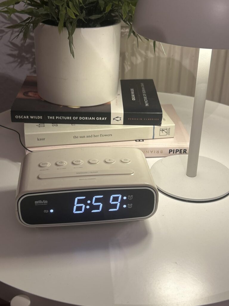 Le radio-réveil posé sur une table de chevet, à côté d'une lampe, de livres et d'une plante. L'heure affichée est 6h59.