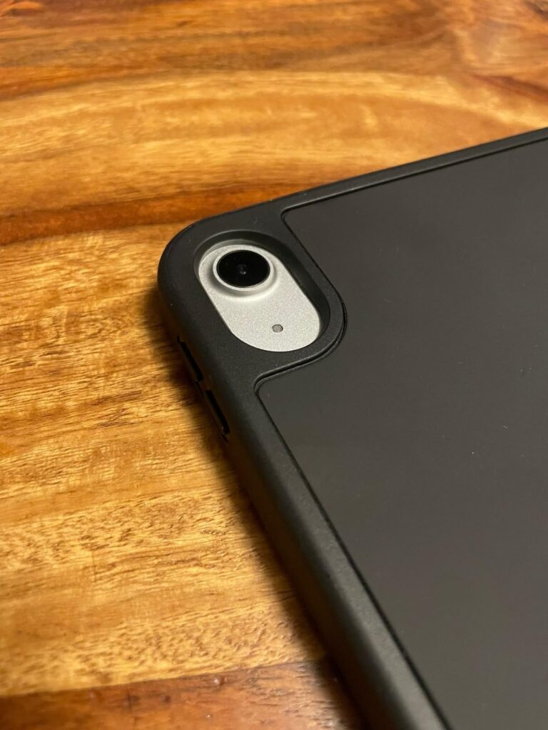 Gros plan sur le coin supérieur gauche de l'iPad avec la coque noire, posé sur une table en bois. Le module caméra est claire...