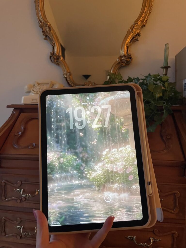 Vue de face de l'iPad en situation, avec son étui couleur crème, tenu par une main. L'écran de l'iPad affiche un paysage avec...