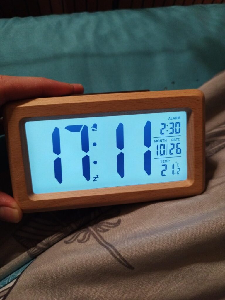 Vue de face du réveil dans une main, affichage lumineux bleu, heure 17:11, alarme, date et température 21°C.