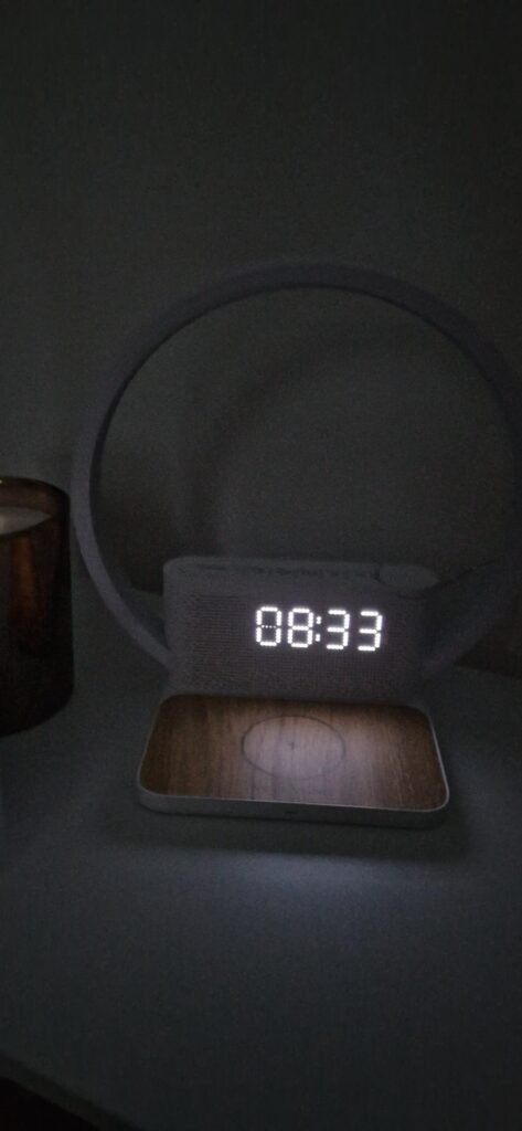 Réveil-chargeur avec lampe d'ambiance allumée, posé sur une surface sombre, devant un mur clair, vu d'un angle légèrement dif...