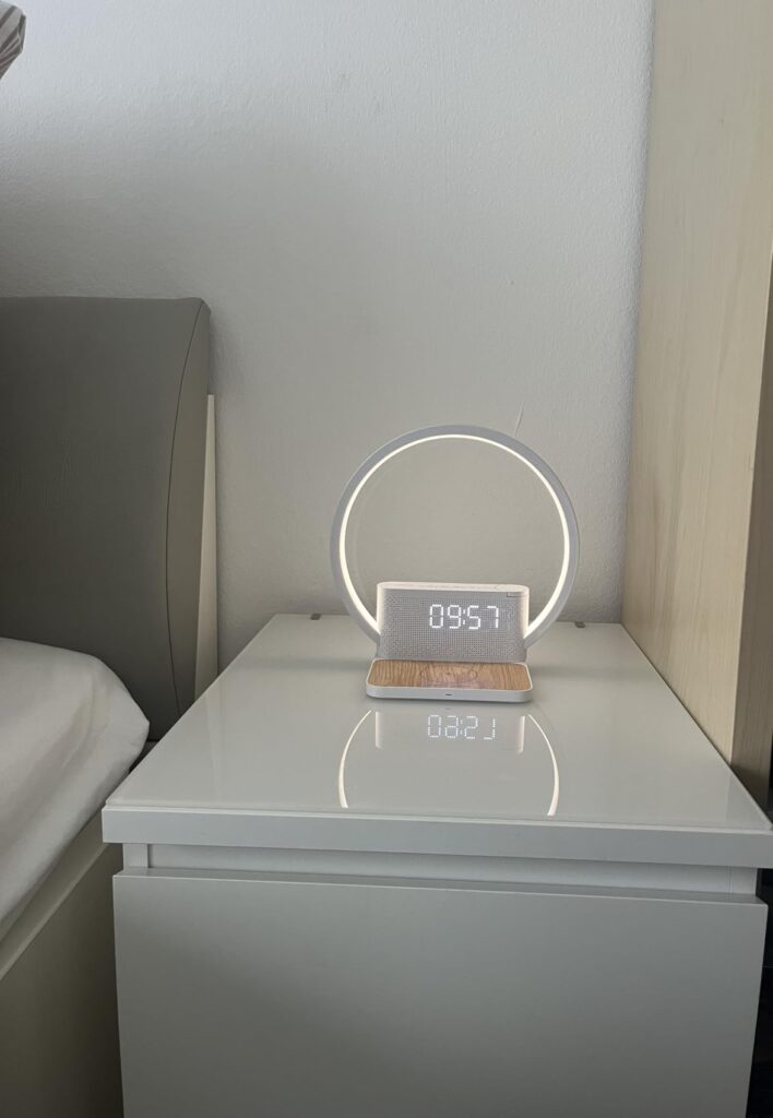 Réveil-chargeur avec lampe d'ambiance allumée, posé sur une table de chevet blanche dans une chambre.