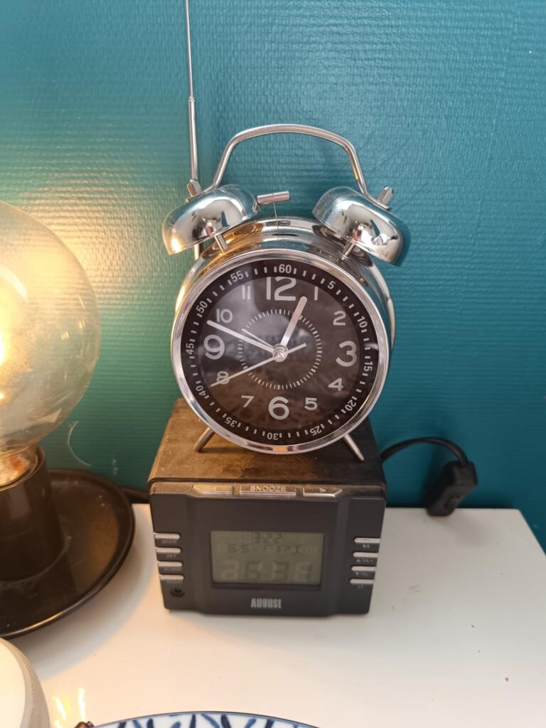 Un réveil à double clochette, de couleur argentée, posé sur une petite radio, sur une table de nuit. Derrière, un mur bleu tu...