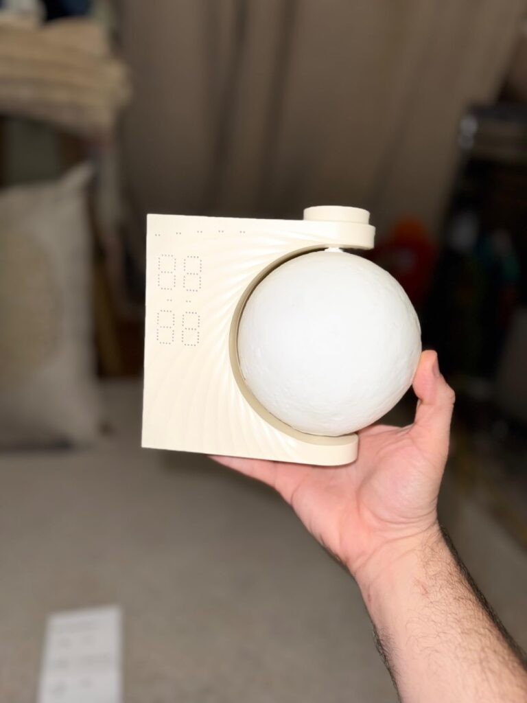 Un homme tient le réveil-lumière dans sa main. La lune est blanche et éteinte. Le réveil est en plastique blanc cassé. Le fon...