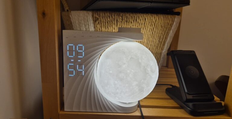 Le réveil-lumière en forme de lune dans une chambre, posé sur une étagère en bois. L'affichage de l'heure est visible : 09:54...