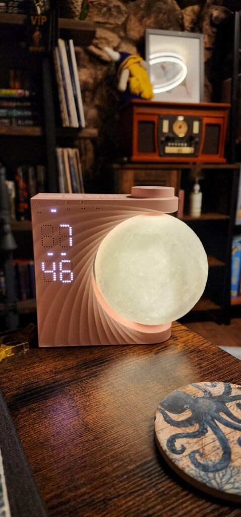 Le réveil-lumière en forme de lune dans une chambre, posé sur une table en bois. L'affichage de l'heure est visible : 7:46. L...