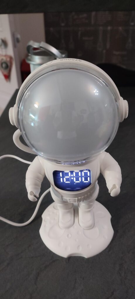 Le projecteur astronaute est allumé et affiche l'heure '12:00' sur son torse. Il est posé sur une surface sombre avec des câb...