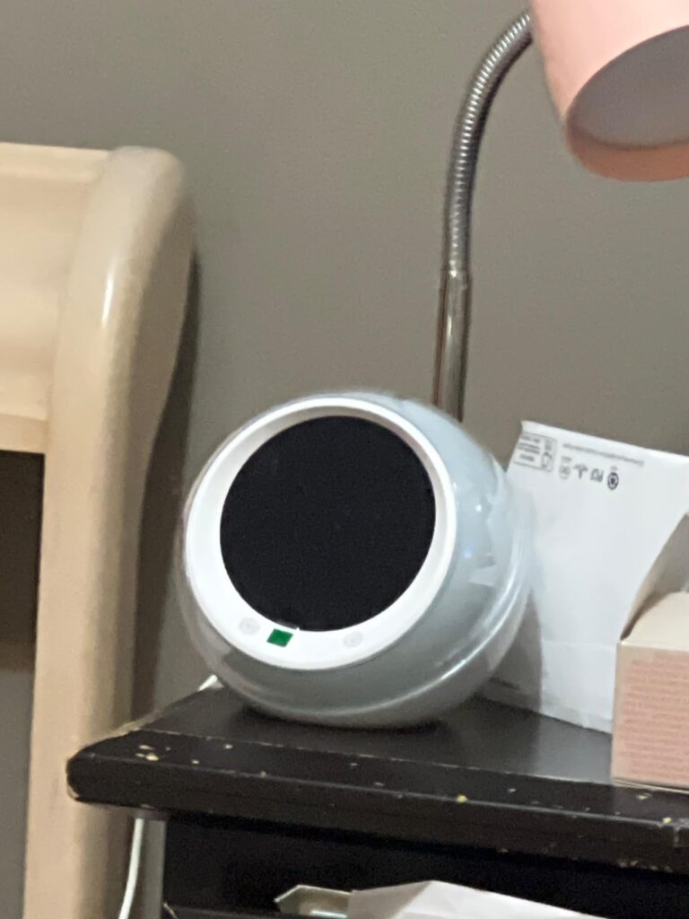 Vue de côté du réveil posé sur une table de chevet, avec un écran éteint et une petite lumière verte allumée. Le réveil est à...
