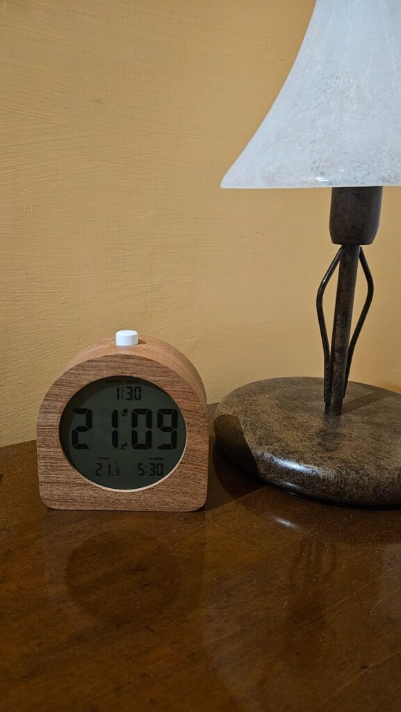 Le même réveil que l'image précédente, posé sur une table en bois à côté d'une lampe de chevet.