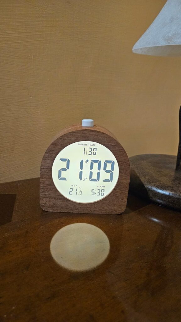 Le réveil avec l'écran allumé (rétro-éclairé), posé sur une table en bois à côté d'une lampe de chevet. Le visuel 1 montre la...