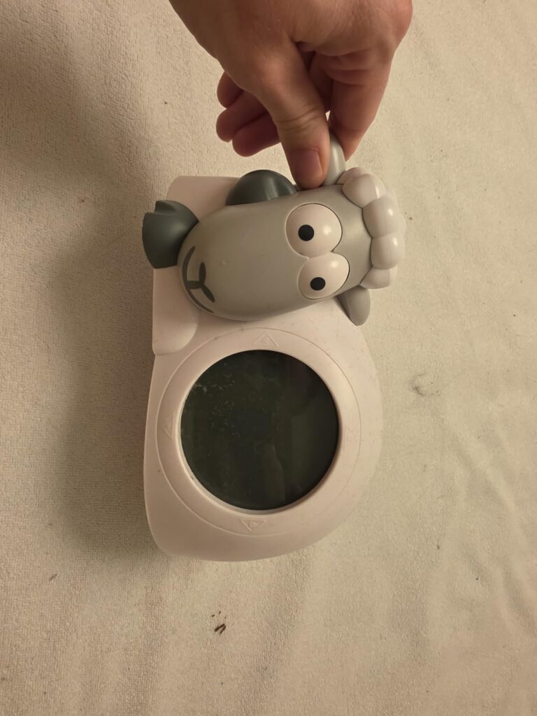 Réveil de jour pour enfant en forme de mouton, vu de face, tenu par la main. Le visuel 1 montre le même objet sous un angle l...