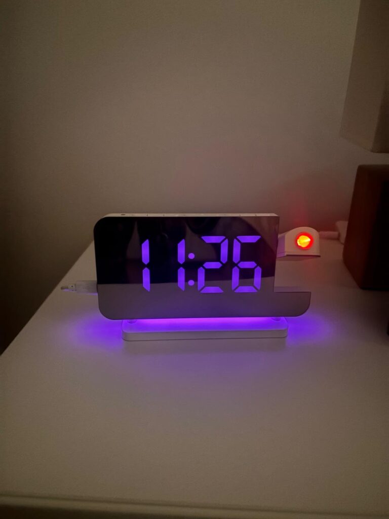 Le réveil sur une table de chevet, l'heure est affichée en violet et la base est éclairée en violet également. On aperçoit un...