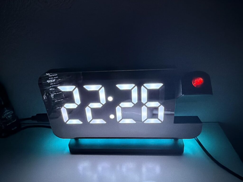Le réveil sur une table de nuit, l'heure est affichée en blanc et la base est éclairée en bleu ciel. On aperçoit le projecteu...