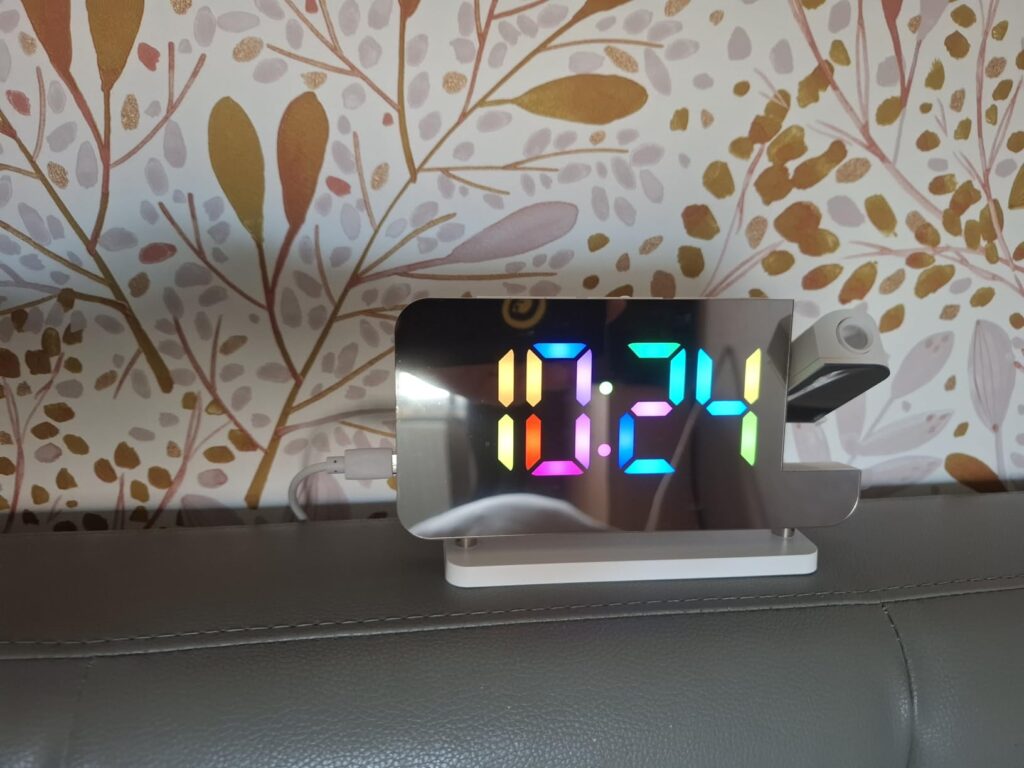 Le réveil sur un meuble, l'heure est affichée en plusieurs couleurs et la base n'est pas éclairée. On aperçoit un mur avec un...