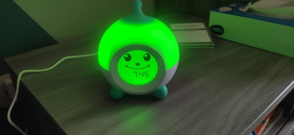 Le réveil est posé sur une table de chevet, avec l'écran affichant l'heure et l'éclairage vert activé. L'image est similaire...