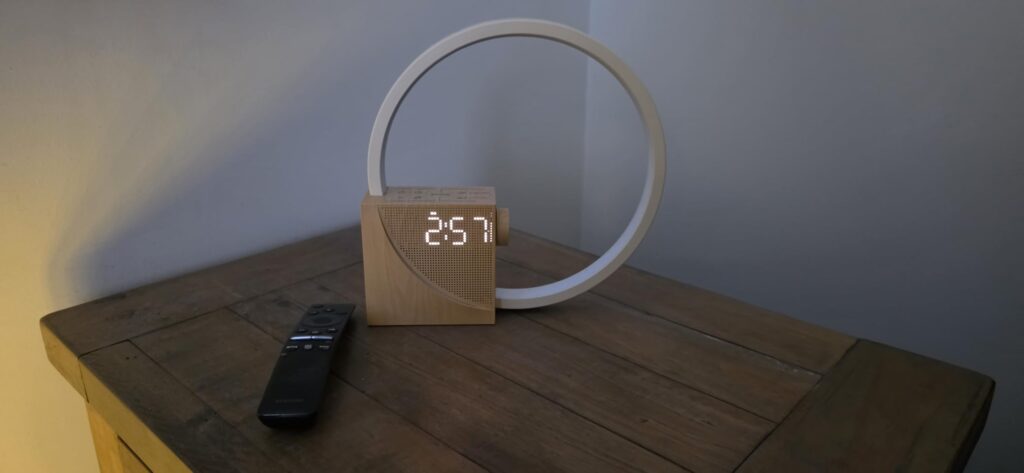 Réveil lumineux sur une table de chevet, allumé, heure affichée à 2h57. Une télécommande est visible à côté du réveil.