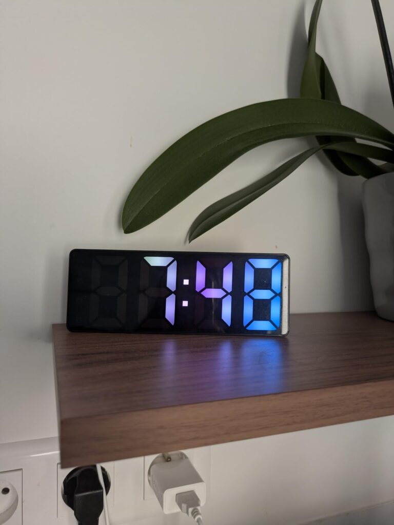 L'horloge dans un salon, posée sur une étagère en bois. Elle affiche 7:48 avec des chiffres multicolores.