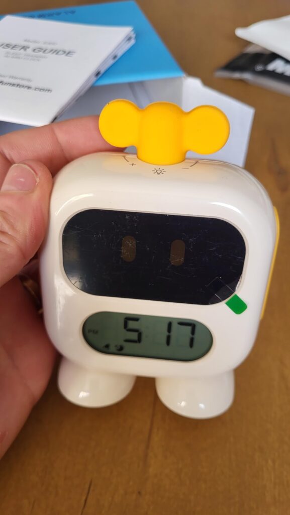Réveil robot tenu dans la main. L'écran du haut est noir, l'écran du bas affiche l'heure en vert. Même image que la 1, angle...