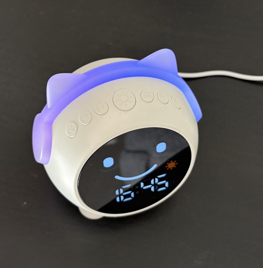 Réveil pour enfant blanc avec oreilles de chat, éclairées en violet. Le réveil est posé sur une surface sombre et l'heure est...