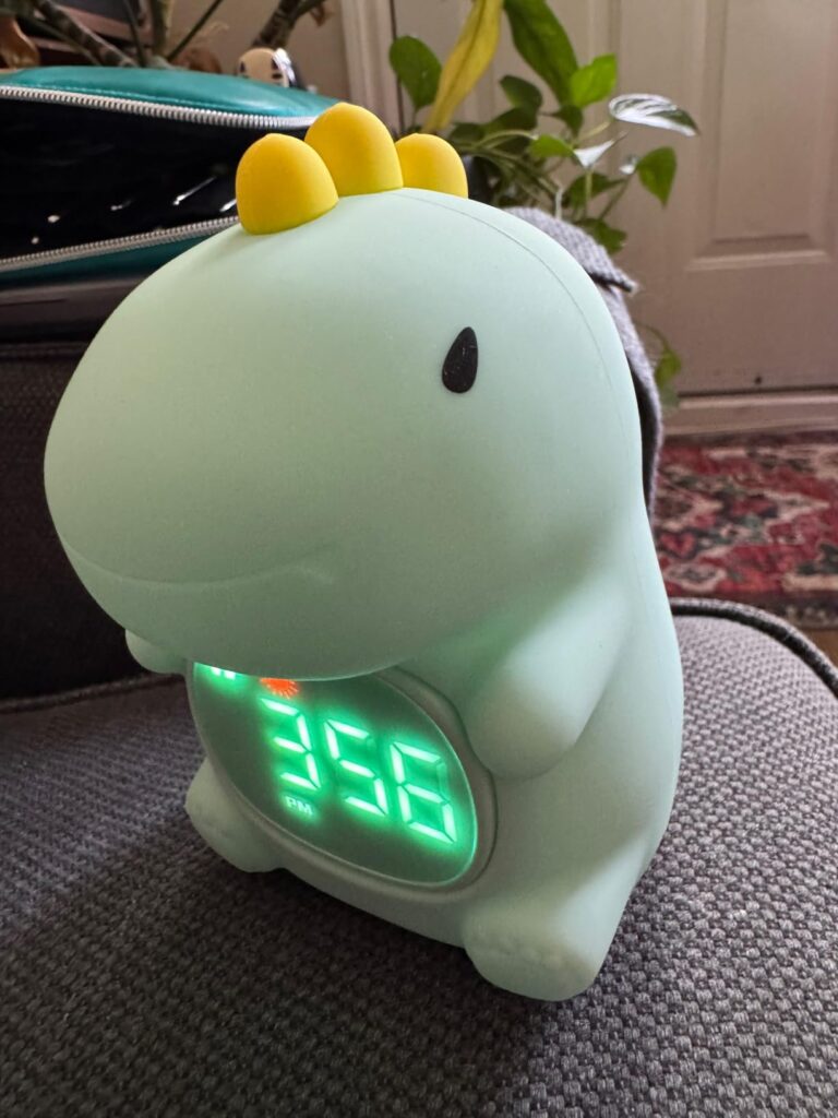 Le réveil dinosaure est posé sur un canapé, l'écran affiche l'heure. Le produit est de couleur verte et jaune. Une plante est...