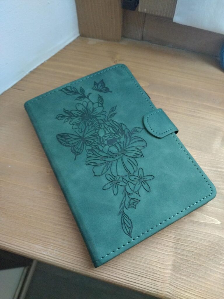 Housse pour liseuse, couleur vert clair, vue de dessus en angle sur un fond en bois. Motif de fleurs et papillons gravé sur l...