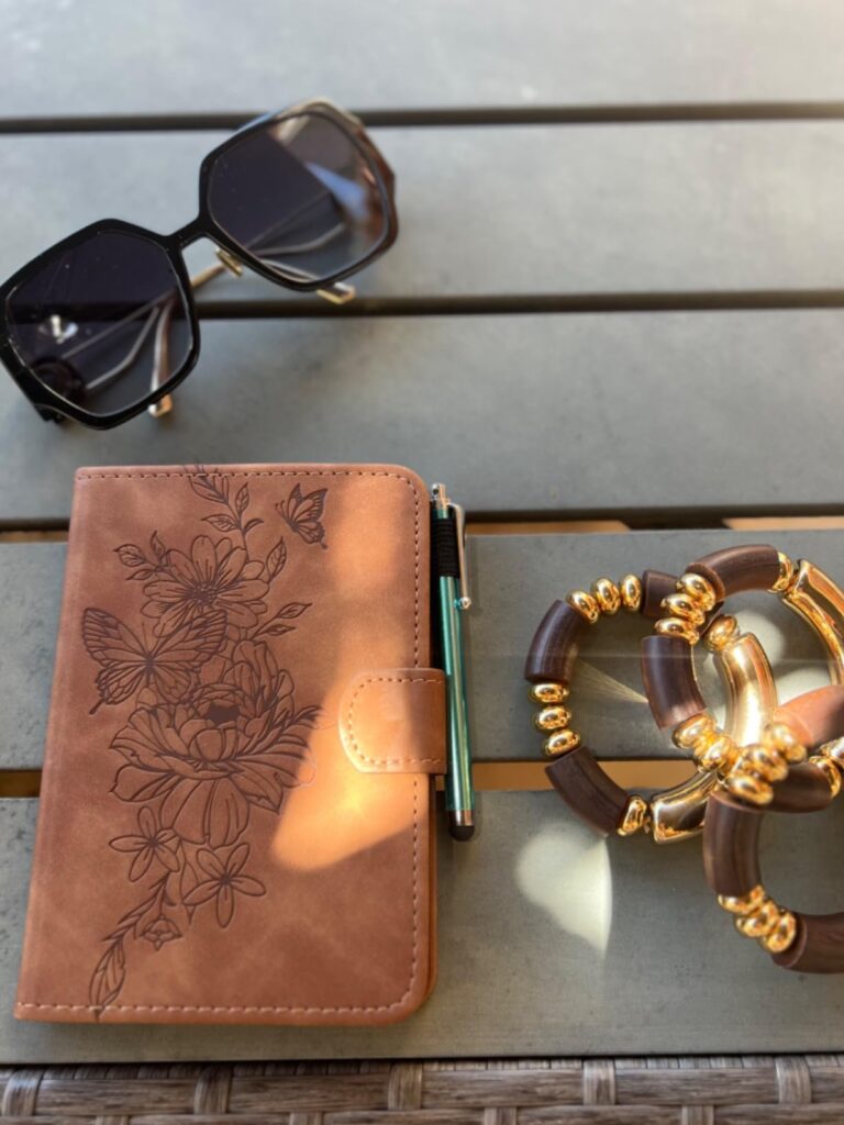 Housse pour liseuse, couleur marron, posée sur une table d'extérieur avec des lunettes de soleil et un bracelet. Un stylo est...