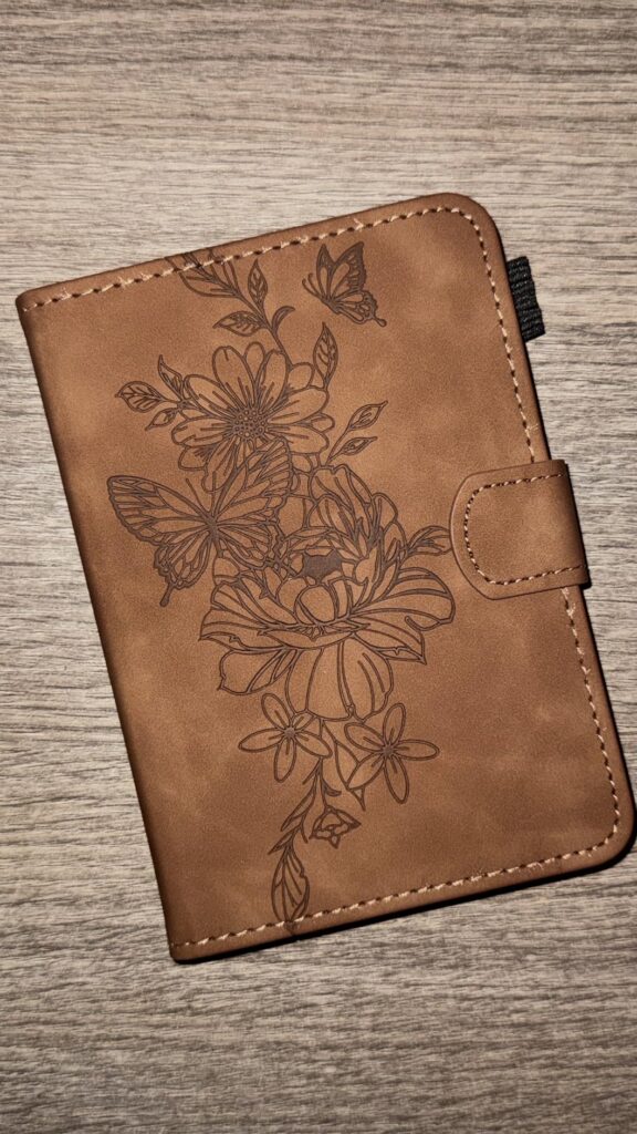 Housse pour liseuse, couleur marron, vue de dessus sur un fond en bois. Motif de fleurs et papillons gravé sur le devant.