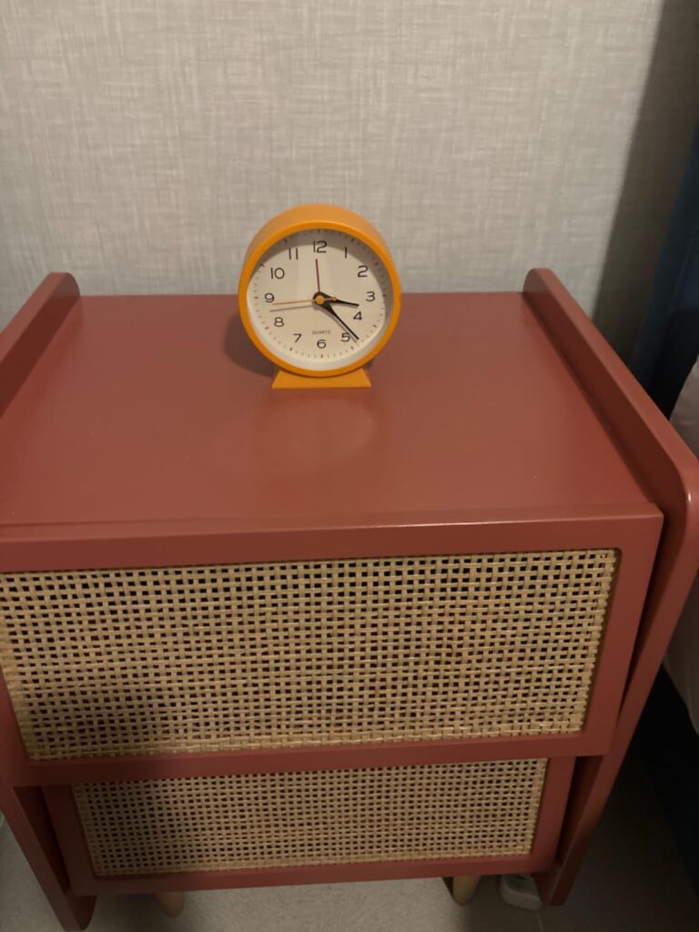 Un réveil jaune sur une table de chevet orange avec des tiroirs en rotin