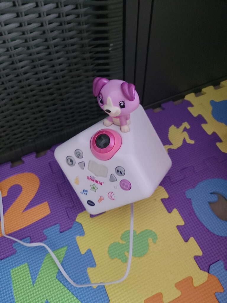 Le conteur d'histoire VTech Story'Clock avec une figurine de chien, posé sur un tapis de jeu pour enfant, vue de côté.