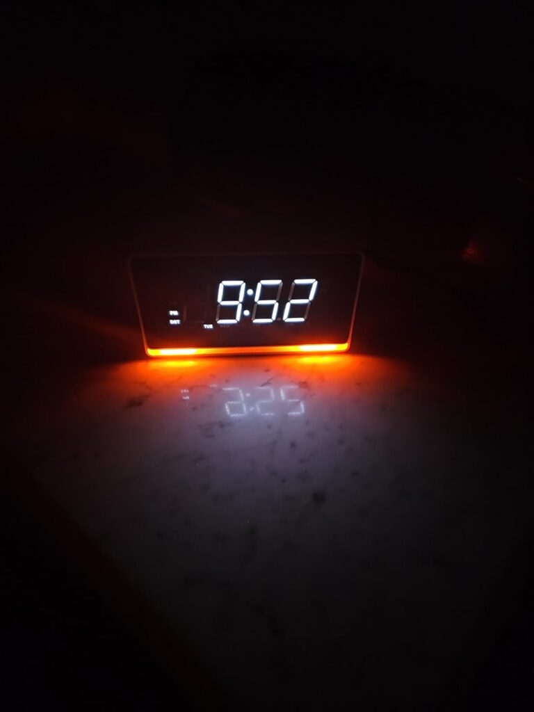 Vue de face du radio-réveil ITOMA dans l'obscurité. L'écran affiche 9:52 et le socle est éclairé en orange, se reflétant sur...