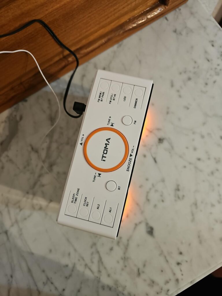 Vue de dessus du radio-réveil ITOMA posé sur une table de chevet, avec les commandes visibles. Le socle est éclairé en orange...