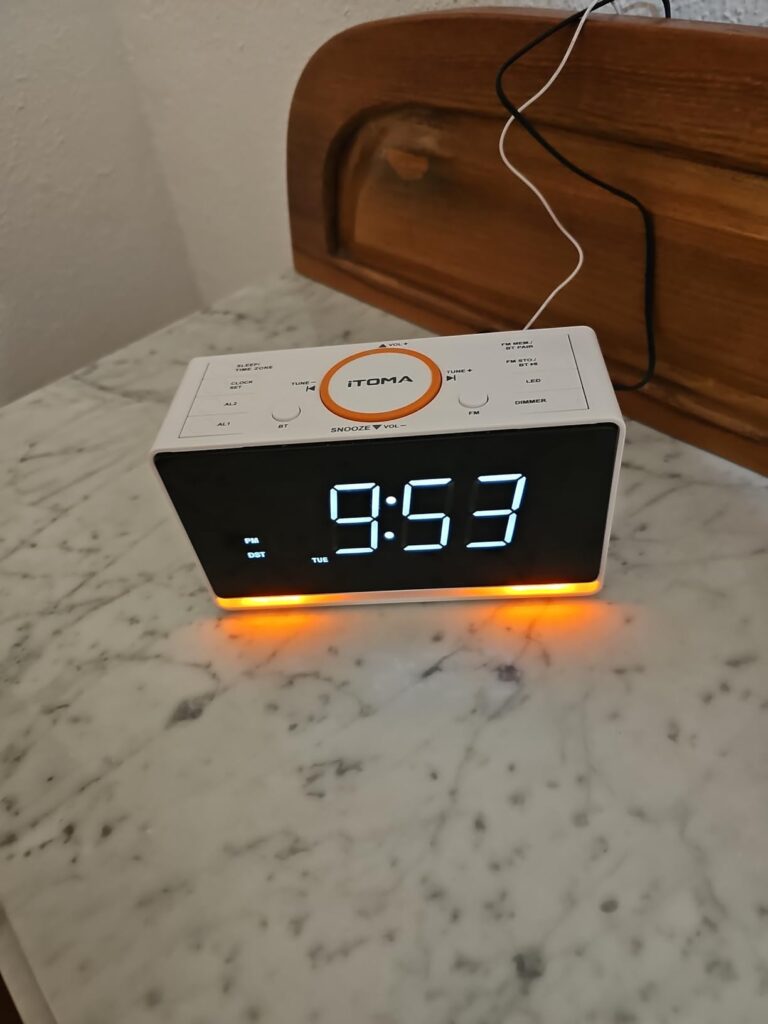 Vue de face du radio-réveil ITOMA posé sur une table de chevet, similaire à l'image précédente. Le socle est éclairé en orang...