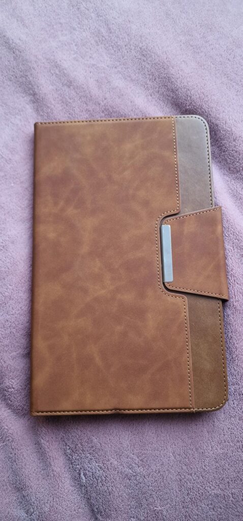 L'étui de protection marron clair pour tablette, fermé, vu de face, posé sur une couverture violette. On voit la languette de...