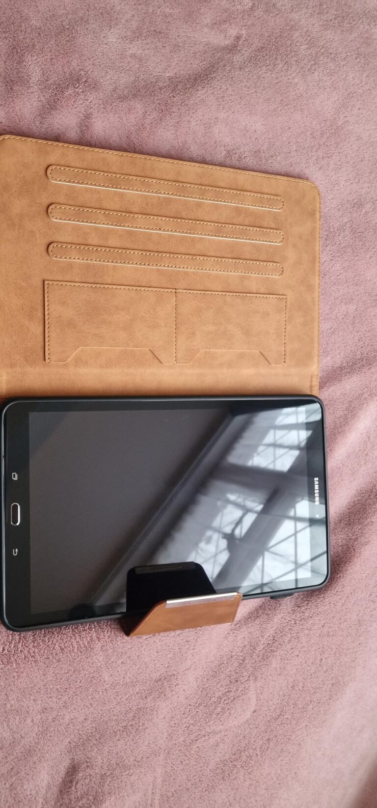 Une tablette noire est insérée dans l'étui marron clair, ouverte en mode support, posée sur une couverture violette. L'intéri...