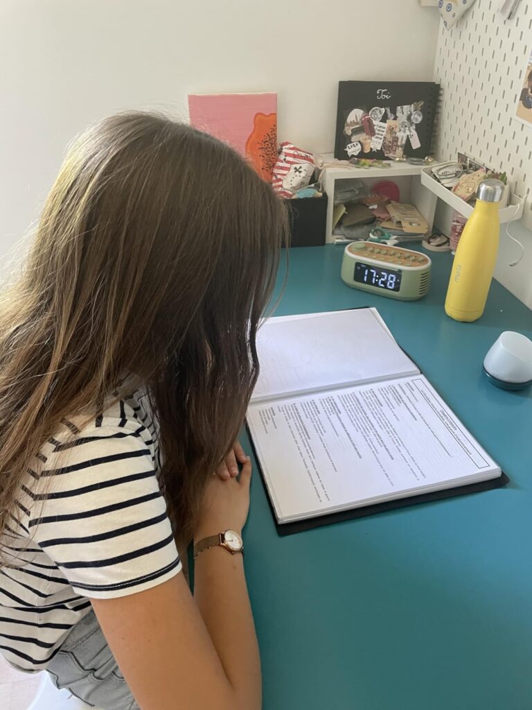 Vue d'une jeune femme de dos, assise à son bureau, le réveil est posé sur le bureau à côté d'une gourde jaune et d'un cahier...