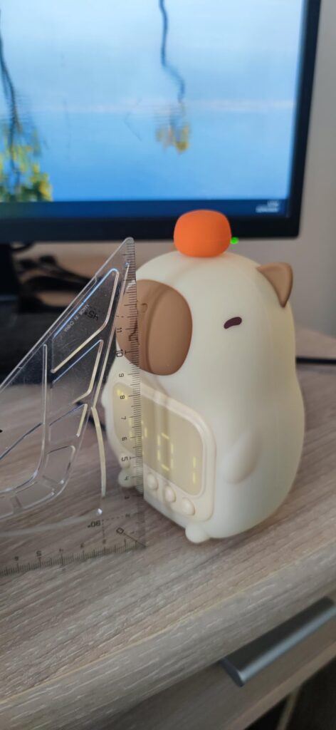 Le réveil en forme de capybara est posé sur un bureau, à côté d'un écran. Le support arrière est en acrylique transparent.