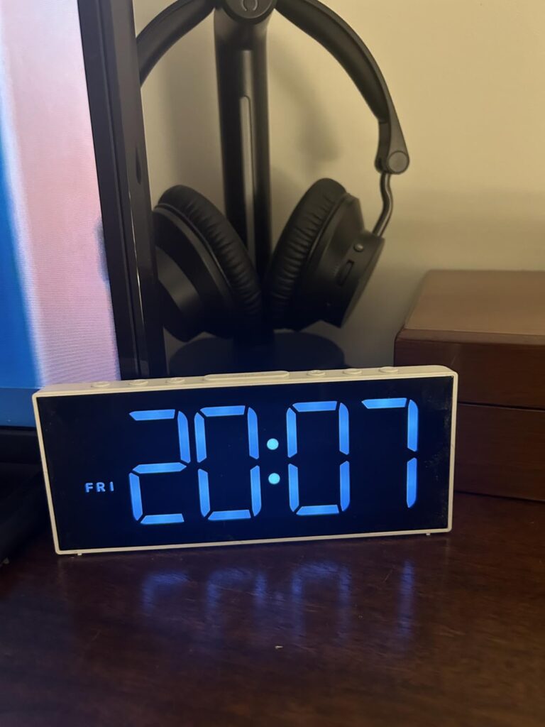 Le réveil sur une table de chevet, affichant l'heure et le jour de la semaine, à côté d'un casque audio posé sur un support.