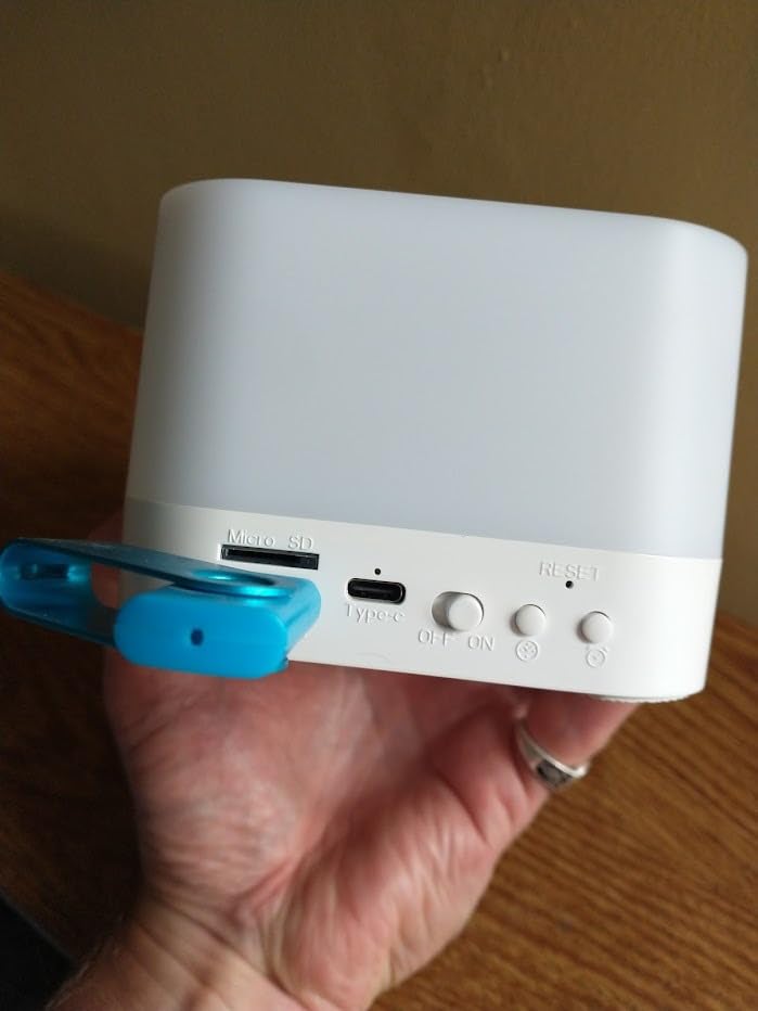 Une main tient le réveil, gros plan sur la face arrière avec le port USB, le port carte SD et les boutons de réglage. Une clé...